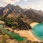 Dramatic coastal landscape in Oman featuring rugged mountains, turquoise waters, sandy beaches and a traditional dhow boat.