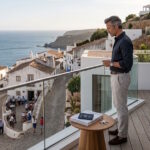 A man standing on a modern luxury villa terrace with a glass railing, looking down at a traditional European coastal village street by the sea.