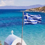 Greek flag overlooking the Aegean Sea with a traditional white chapel on the Greek coastline