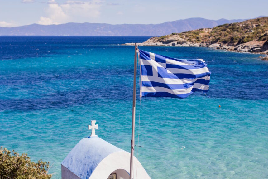 Greek flag overlooking the Aegean Sea with a traditional white chapel on the Greek coastline